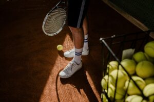 A tennis player stands on a clay court, showing sneakers, racket, and tennis balls in sunlight.