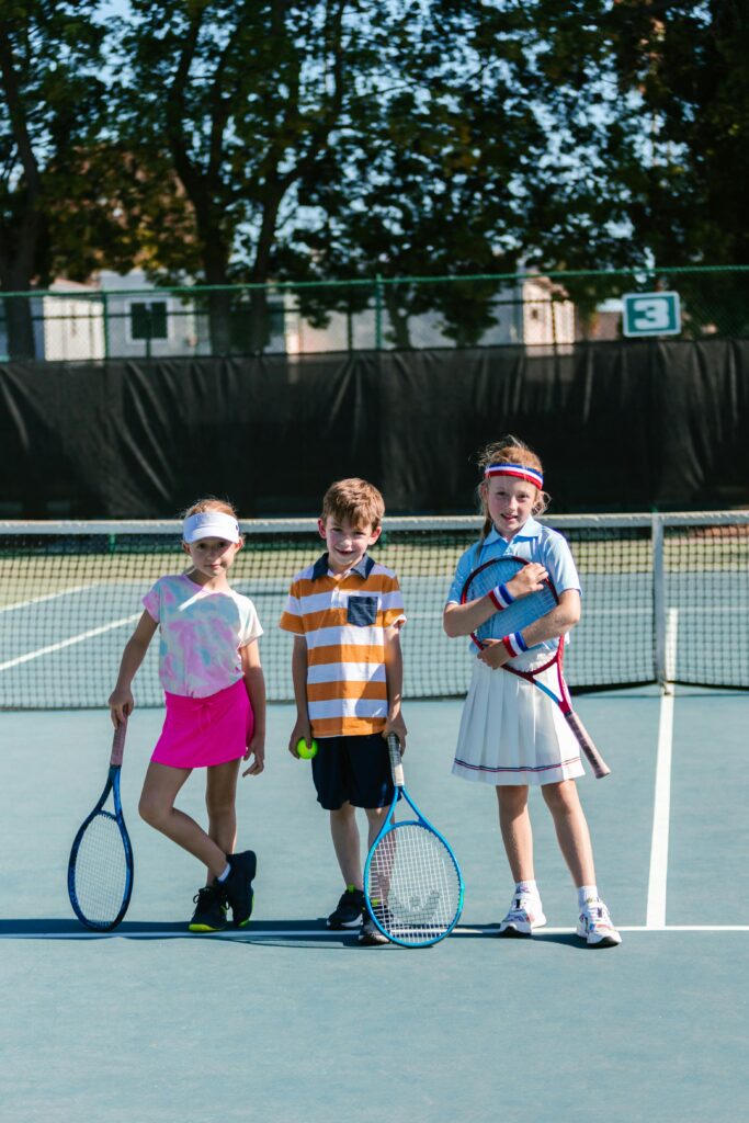 Three children having fun on an outdoor tennis court during practice, wearing sporty outfits.